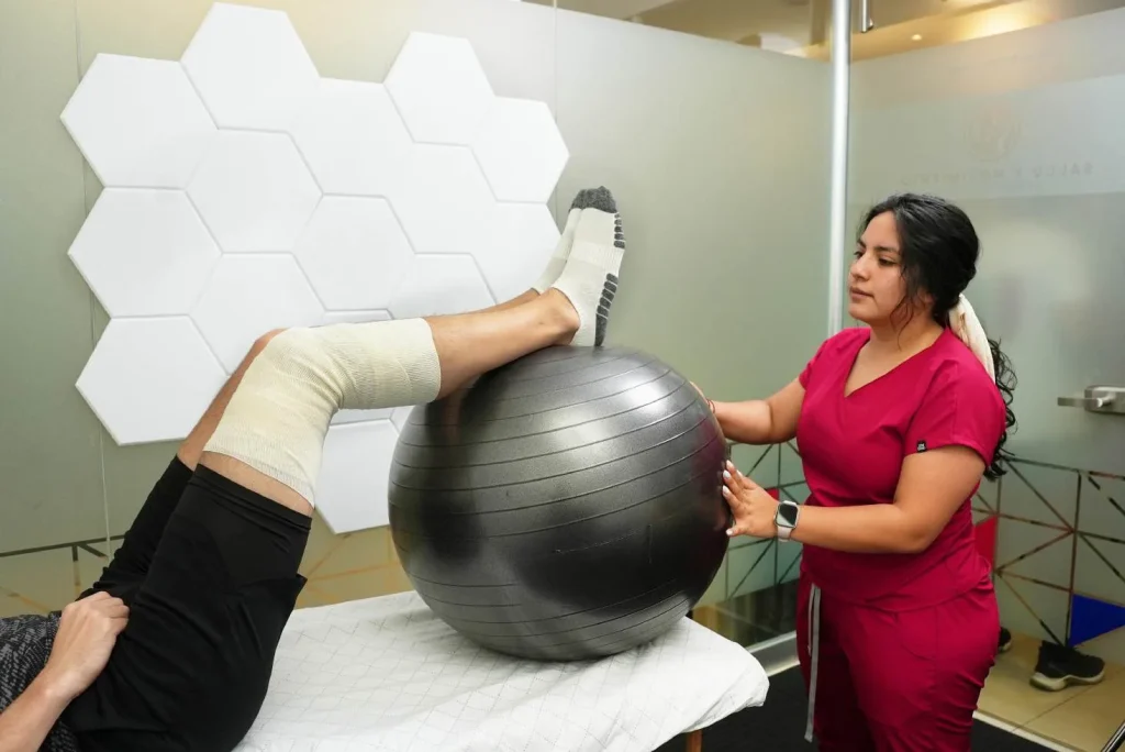 Ejercicio de rehabilitación con pelota terapéutica bajo las piernas, guiado por la fisioterapeuta